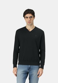 Maglione nero a scollo a V realizzato in tessuto liscio, con polsini e orlo a coste, indossato sopra jeans azzurri con un dettaglio logo discreto sul petto.