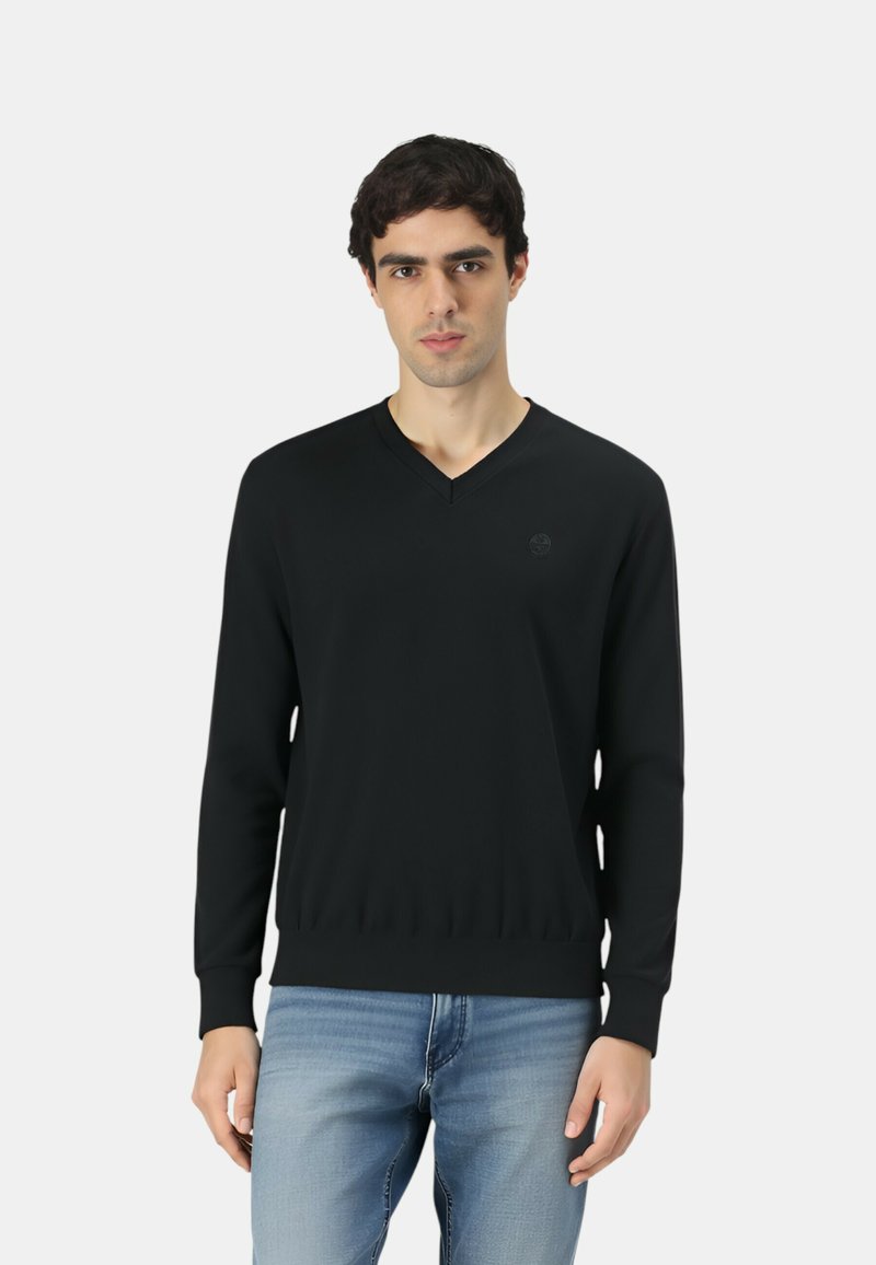 Maglione nero a scollo a V realizzato in tessuto liscio, con polsini e orlo a coste, indossato sopra jeans azzurri con un dettaglio logo discreto sul petto.