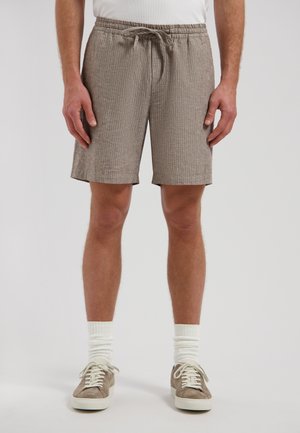 Mann trägt beige strukturierte Shorts mit Kordelzug, weiße Socken und beige Sneakers und steht vor einem einfarbigen Hintergrund.