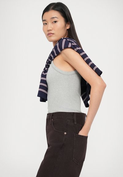 Szürkés ujjatlan top bordás textúrával, sötétbarna farmernadrággal párosítva. Egy tengerészkék csíkos pulóver lóg a vállakon.