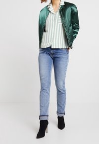 Kvinna klädd i en grön satinbomberjacka över en randig skjorta, ihop med ljusblå jeans och svarta klackboots, stående med självförtroende.