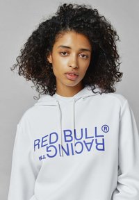 Sudadera blanca con el texto azul "RED BULL RACING™" impreso verticalmente. Cuenta con una capucha con cordones y una tela suave con una textura lisa.