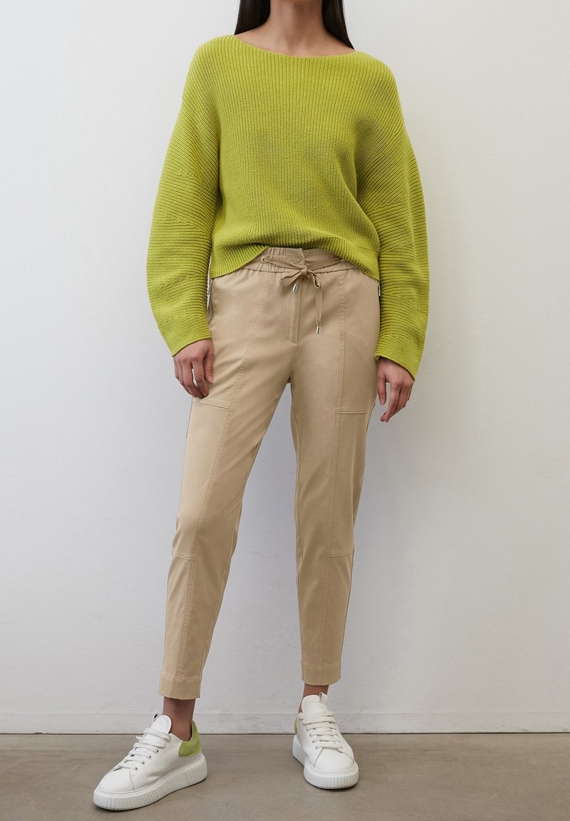 Femme portant un pull en maille vert citron, un pantalon beige avec cordon et des baskets blanches avec un détail vert au talon, debout contre un mur uni.