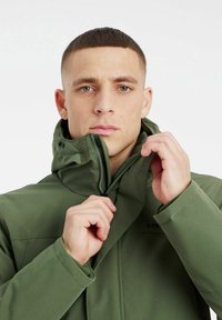 Veste zippée verte avec un col haut et une capuche ajustable. Présente une texture lisse et un logo sur la poitrine. Le modèle ajuste la fermeture éclair.
