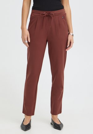 Femme portant un pantalon à cordon couleur rouille avec plis à l'avant et des ballerines noires pointues, debout devant un fond clair uni.