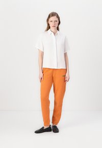 Camicia bianca con maniche corte e bottoni, pantaloni della tuta arancioni con testo verde, e scarpe nere basse, in piedi contro uno sfondo neutro.