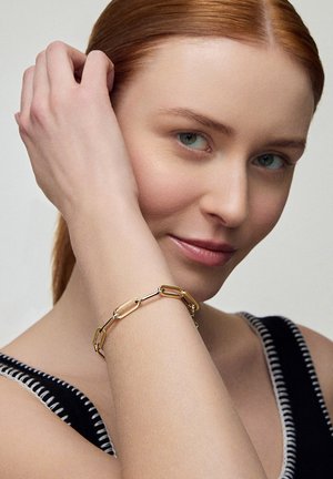Bransoletka metalowa w kolorze złotym, z wydłużonymi, splatanymi ogniwami. Noszona na nadgarstku, prezentująca gładkie powierzchnie i bezszwowy design.