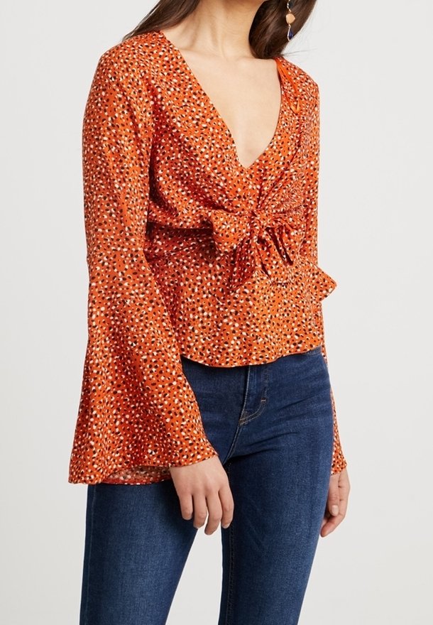 Blouse orange avec un décolleté en V profond, détail noué à la taille et manches évasées. Présente un motif à pois de différentes couleurs.