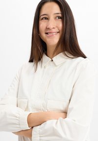 Wit overhemd met lange mouwen, geplooide manchetten en twee voorzakken. Glad textuur en lichtgewicht stof.