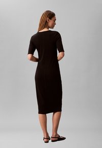 Femeie stând cu fața spre spate, purtând o rochie neagră până la genunchi cu mâneci trei sferturi și pantofi negri fără toc, pe un fundal gri simplu.