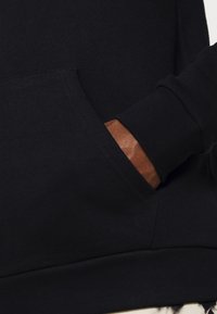 Juodasis džemperis su kengūros kišene ir rudos odos akcentais ant rankogalių. Audinys atrodo minkštas su lygia tekstūra.