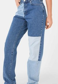 Mujer vestida con jeans de patchwork con paneles de denim azul oscuro y claro, con la mano descansando cerca del bolsillo sobre un fondo blanco.