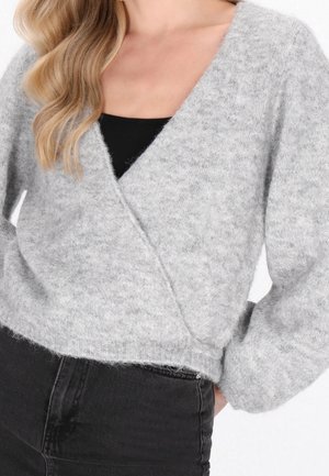 Kobieta nosząca miękki, jasnoszary sweter typu wrap na czarnym topie, połączony z czarnymi dżinsami z wysokim stanem, pokazując tułów i ramiona.
