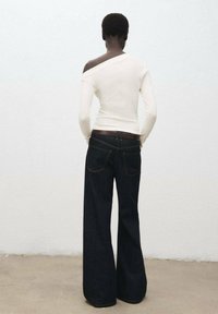 Witte, lange mouw top met een off-the-shoulder ontwerp. Donkerblauwe flared jeans met contrasterende stiksels en een bruine leren riem. Achteraanzicht.