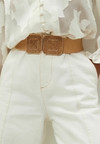 Geflochtener, tanfarbener Gürtel mit quadratischen, strukturierten goldfarbenen Schnallen, getragen über einer weißen Bluse mit Blumenmuster und einer weißen, hoch taillierten Hose mit sichtbaren Nähten.