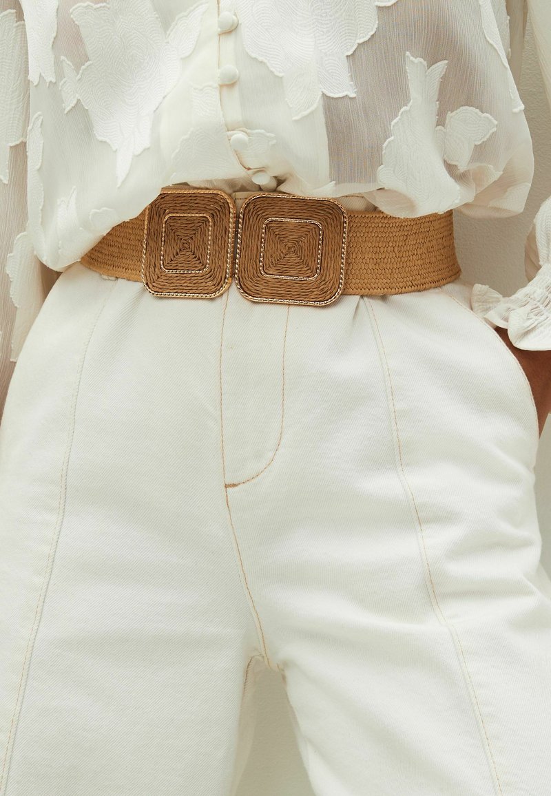 Geflochtener, tanfarbener Gürtel mit quadratischen, strukturierten goldfarbenen Schnallen, getragen über einer weißen Bluse mit Blumenmuster und einer weißen, hoch taillierten Hose mit sichtbaren Nähten.