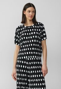 Zwarte geplooide top met grote witte stippen, ronde halslijn, korte mouwen en een relaxed fit. De stof heeft een gestructureerde textuur.