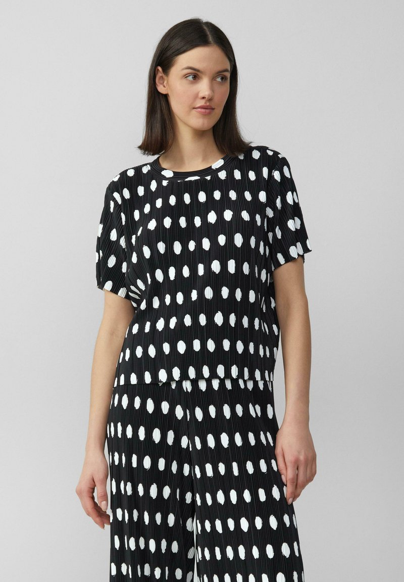 Zwarte geplooide top met grote witte stippen, ronde halslijn, korte mouwen en een relaxed fit. De stof heeft een gestructureerde textuur.