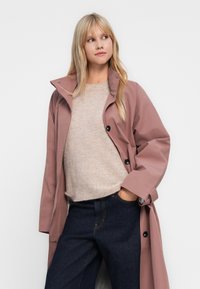 Femme blonde portant un manteau rose poussiéreux sur un pull beige et un jean bleu foncé, se tenant avec une main dans sa poche, regardant à droite.