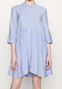 Světle modrá košilová šaty s tříčtvrtečními rukávy, nařaseným pasem a měkkou, hladkou texturou. Má kulatý výstřih a mírný lesk.