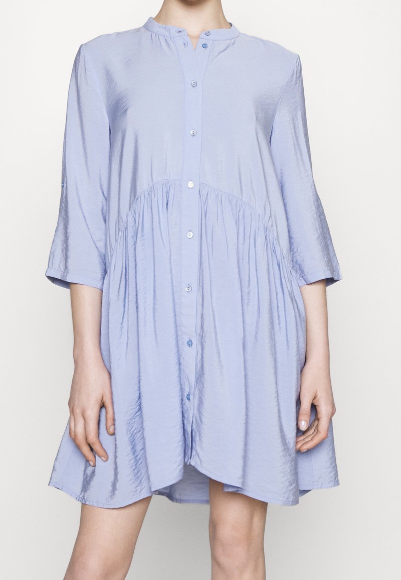 Světle modrá košilová šaty s tříčtvrtečními rukávy, nařaseným pasem a měkkou, hladkou texturou. Má kulatý výstřih a mírný lesk.