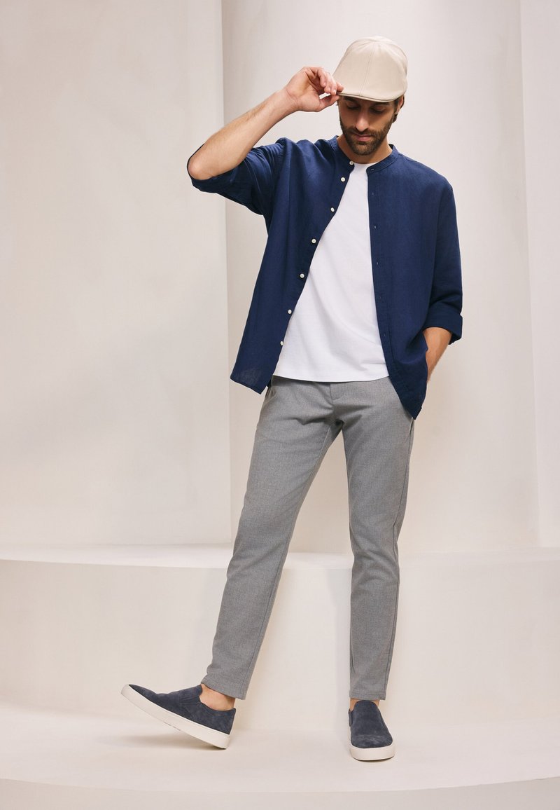 Marineblauw knoopshirt over een wit T-shirt, grijze tapered broek en marineblauwe instappers. Beige pet, minimalistisch ontwerp, gladde texturen.