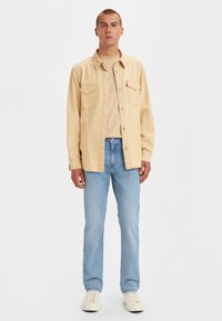 Camisa de botón beige clara, con dos bolsillos en el pecho, llevada sobre una camiseta beige. Jeans azul claro y zapatillas blancas completan el atuendo.