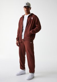 Felpa marrone con zip e logo bianco, abbinata a pantaloni della tuta coordinati e sneakers bianche. Il modello indossa un berretto grigio con accenti blu navy.