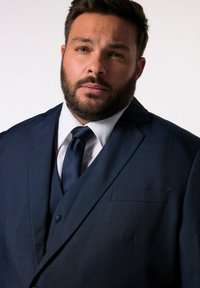Granatowy garnitur z zapięciem na jeden guzik, klapami w kształcie notcha i teksturowanym materiałem; połączony z białą koszulą i jednolitą granatową krawatem.