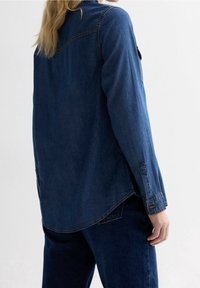 Person som bär en mörkblå denimskjorta och jeans, visad från ryggen mot en slät vit bakgrund.