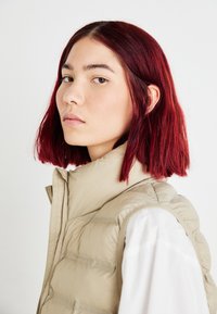 Chaleco acolchado beige con cuello alto, con costuras horizontales y una textura suave, combinado con una camiseta blanca. Cabello rojo a la altura de los hombros visible.