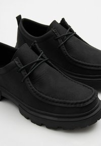 Zapatos de cuero negro con una textura suave, con punta redonda, detalles de costura y suela de goma texturizada. Los cordones se cruzan en la parte delantera.