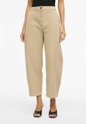 Femme portant un pantalon large taille haute beige assorti à des mules à talons ouvertes noires, debout sur un fond blanc.