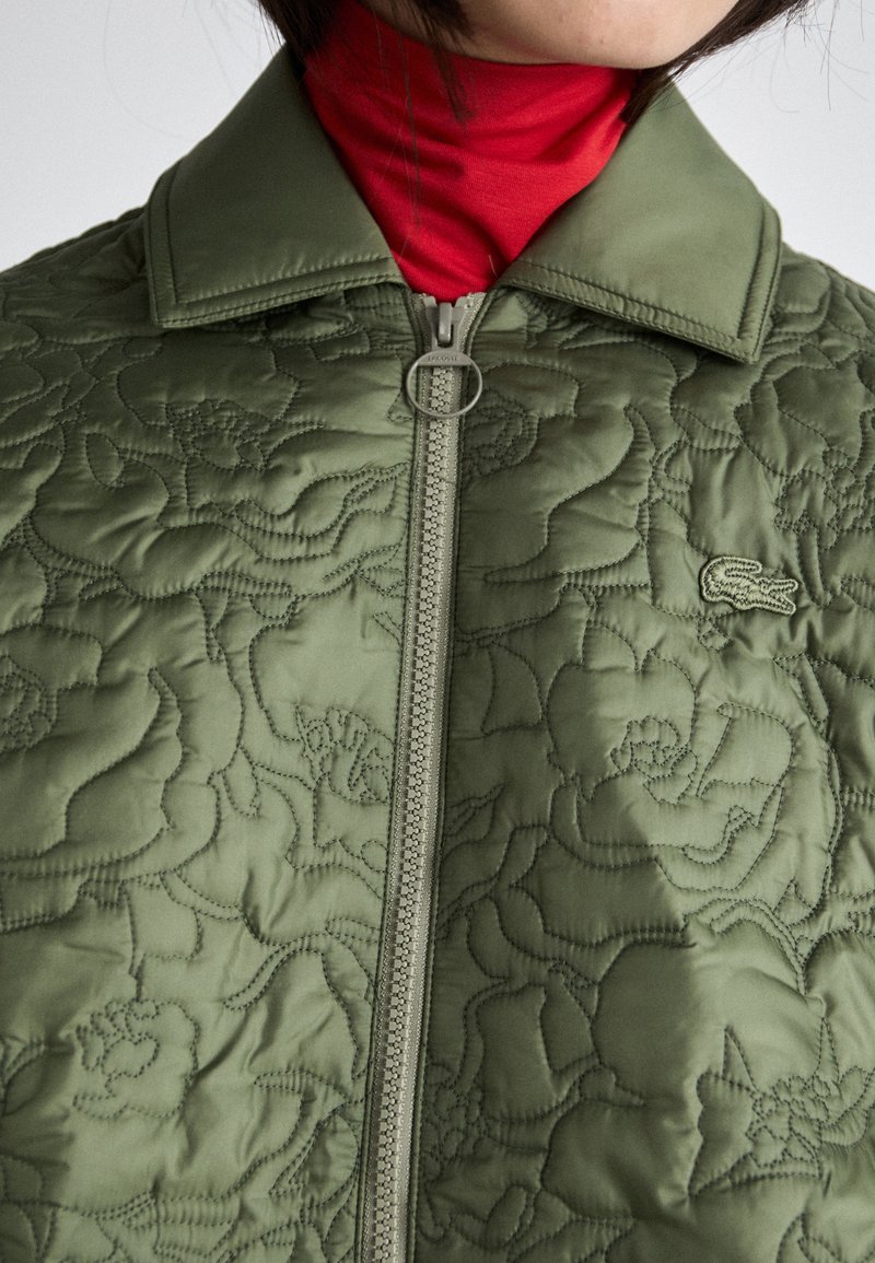 Gepolsterte olivgrüne Jacke mit floralem Muster, Kragen und Frontreißverschluss mit Metallbeschlägen. Darunter getragen wird ein roter Rollkragenpullover.