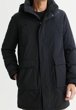 Mężczyzna ubrany w czarną zimową kurtkę z kapturem, z przodu z zamkiem błyskawicznym, zatrzaskami oraz czterema dużymi kieszeniami, ręce częściowo wsunięte do kieszeni.