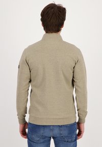 Sudadera color caqui con cuello alto, hecha de un tejido texturizado. Cuenta con puños y dobladillo de canalé, y un parche con el logo en la manga izquierda.