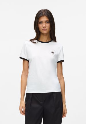 Mujer con camiseta blanca de manga corta con cuello y bordes de las mangas negros, pantalones negros, de pie frente a un fondo blanco.