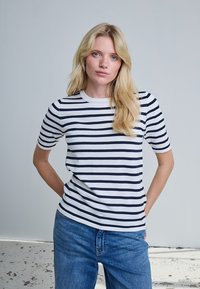 Femme blonde portant un jean bleu et un t-shirt blanc à manches courtes avec des rayures horizontales noires, debout les mains derrière le dos.