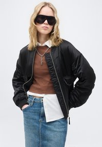 Νεαρή γυναίκα με κυματιστά ξανθά μαλλιά φοράει μαύρα γυαλιά ηλίου μεγάλης διάστασης, μαύρο μπουφάν τύπου μπομπέρ, καφέ πουλόβερ πάνω από λευκό πουκάμισο και μπλε τζιν φούστα.