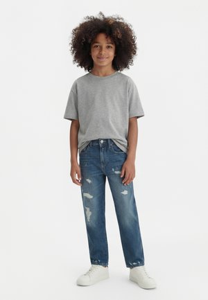 Bērns ar cirtainiem matiem, valkājot pelēku T-kreklu, nodilušas zilas džinsas un baltus sporta apavus, stāvot pret vienkāršu baltu fonu.