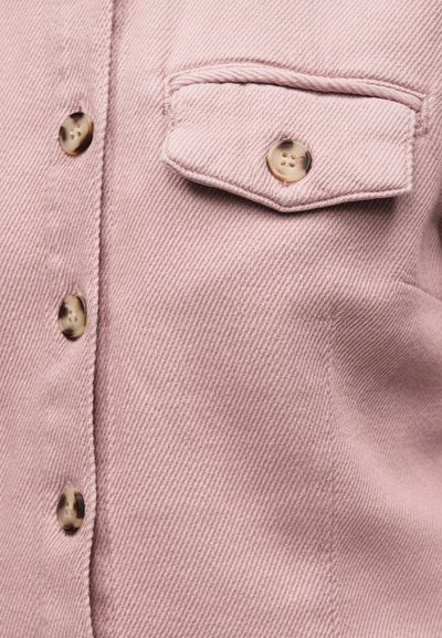 Ροζ υφή υφάσματος με μπεζ μάρμαρα κουμπιά, που δείχνει μια κουμπωτή τσέπη με καπάκι και μέρος από το μπροστινό μέρος ενός ενδύματος με κουμπιά.