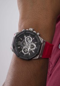 Zilveren octagonale horloge met zwarte wijzerplaat, rode rubberen band en zilveren accenten. Voorzien van drie subwijzers en opvallende uurmarkeringen.