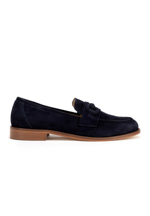 Mocasines de ante azul marino con suela plana marrón, que presentan un detalle de lazo en la parte frontal y un diseño de punta redondeada.
