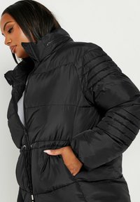 Svart puffjacka med hög krage, dragkedjestängning, ribbade detaljer på ärmarna och dragsko i midjan. Slät, glänsande yta.