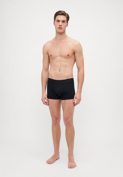 Boxer da uomo neri con una texture liscia e morbida e una vita elasticizzata. Design aderente che migliora il comfort. Il modello è in piedi a piedi nudi su uno sfondo neutro.