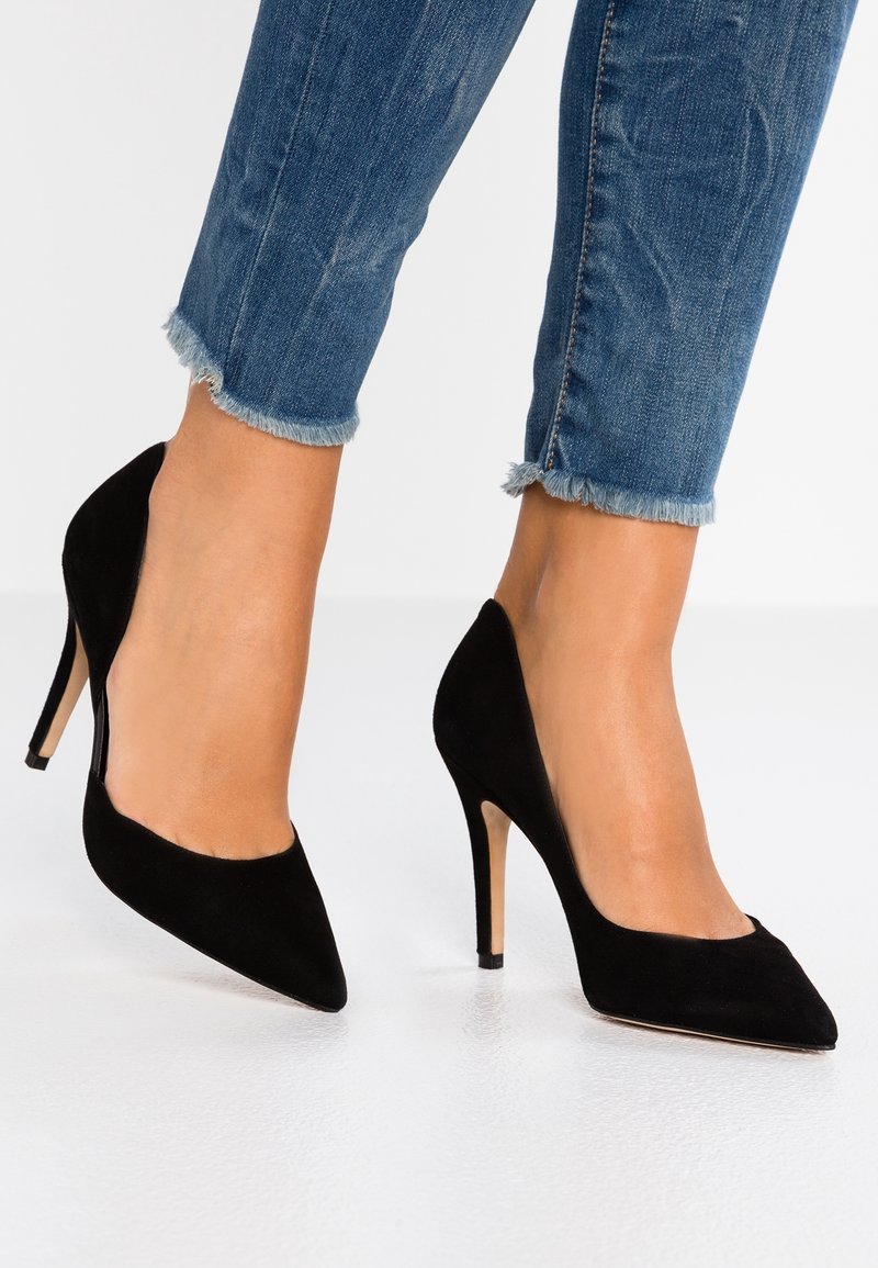Jambes portant un jean bleu écourté avec des ourlets effilochés et des escarpins en daim noir à bout pointu sur un sol blanc.