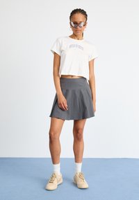 T-shirt cropped blanc avec imprimé "Nike Sports". Jupe plissée grise. Baskets beiges avec lacets. Le modèle pose sur une surface bleu clair.