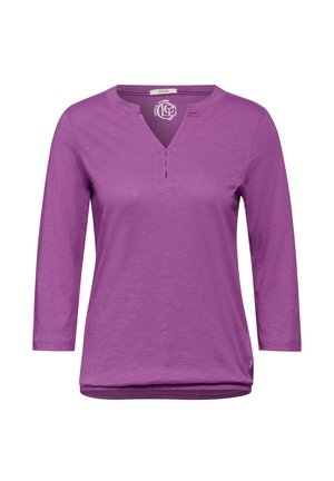 Fioletowa bluzka z długim rękawem, posiadająca lekko dekolt w kształcie V, z krótkim rozcięciem i miękkim, teksturowanym materiałem. Okrągły detal u dołu.