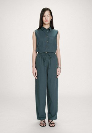 Femme debout, de face, portant un haut sans manches boutonné vert sarcelle foncé et un pantalon ample assorti, ainsi que des sandales noires à lanières.