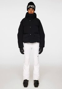 Svart vadderad jacka med tryckknappar, kombinerad med vita skidbyxor med strukturerade ränder. Svarta handskar och mössa med texten "APRÈS".